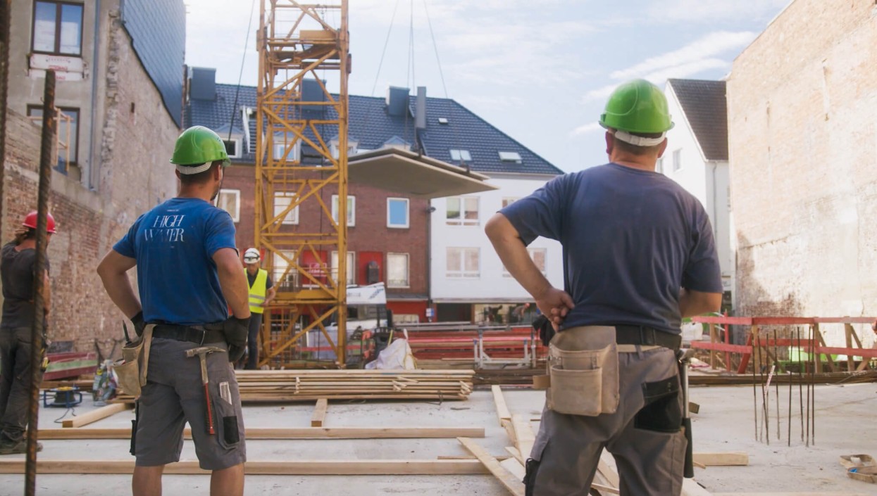 Zwei Personen schauen sich die Baustelle an und nutzen den digitalen Bauablaufplan. Im Hintergrund sind weitere Bauleiter und Poliere auf der Baustelle zu sehen.